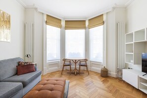 A room with a grey sofa, a round wooden table with chairs, a white TV stand, and a bay window with beige curtains. - GuestReady - Modern Oasis in Kensington (London)