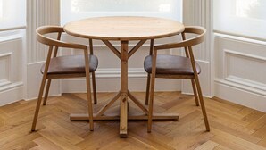 Et rundt træbord med fire stole i et hjørneværelse med store vinduer og beige gardiner.