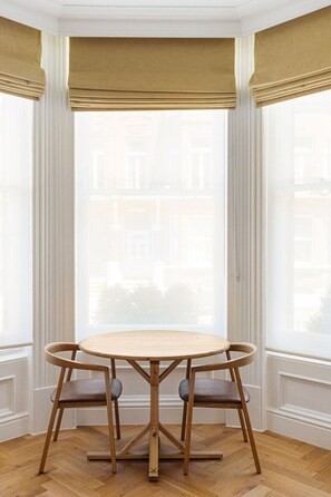 A round wooden table with four chairs in a corner room with large windows and beige curtains. - GuestReady - Modern Oasis in Kensington (London)