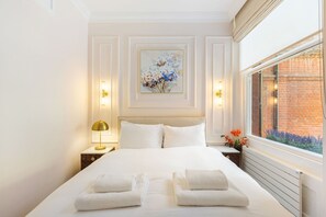 A neatly made bed with white linens and towels, a painting on the wall, and a window with a brick wall view. - GuestReady - Modern Oasis in Kensington (London)