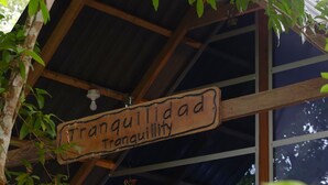 A wooden structure with a sign reading 'Tranquillidad Tranquillity,' a stone wall, and a bed with a white sheet. - Tranquilidad Cabin, Left Hemisphere (San José Del Guaviare)