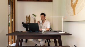 Una persona sentada a una mesa con una computadora portátil, una taza y un vaso de agua. Hay una planta y un objeto decorativo sobre la mesa.