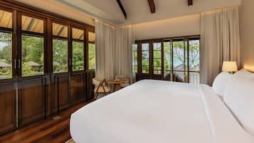 Quarto de hotel com uma cama grande, uma mesinha com cadeiras e amplas janelas que oferecem vista para o verde e um edifício.