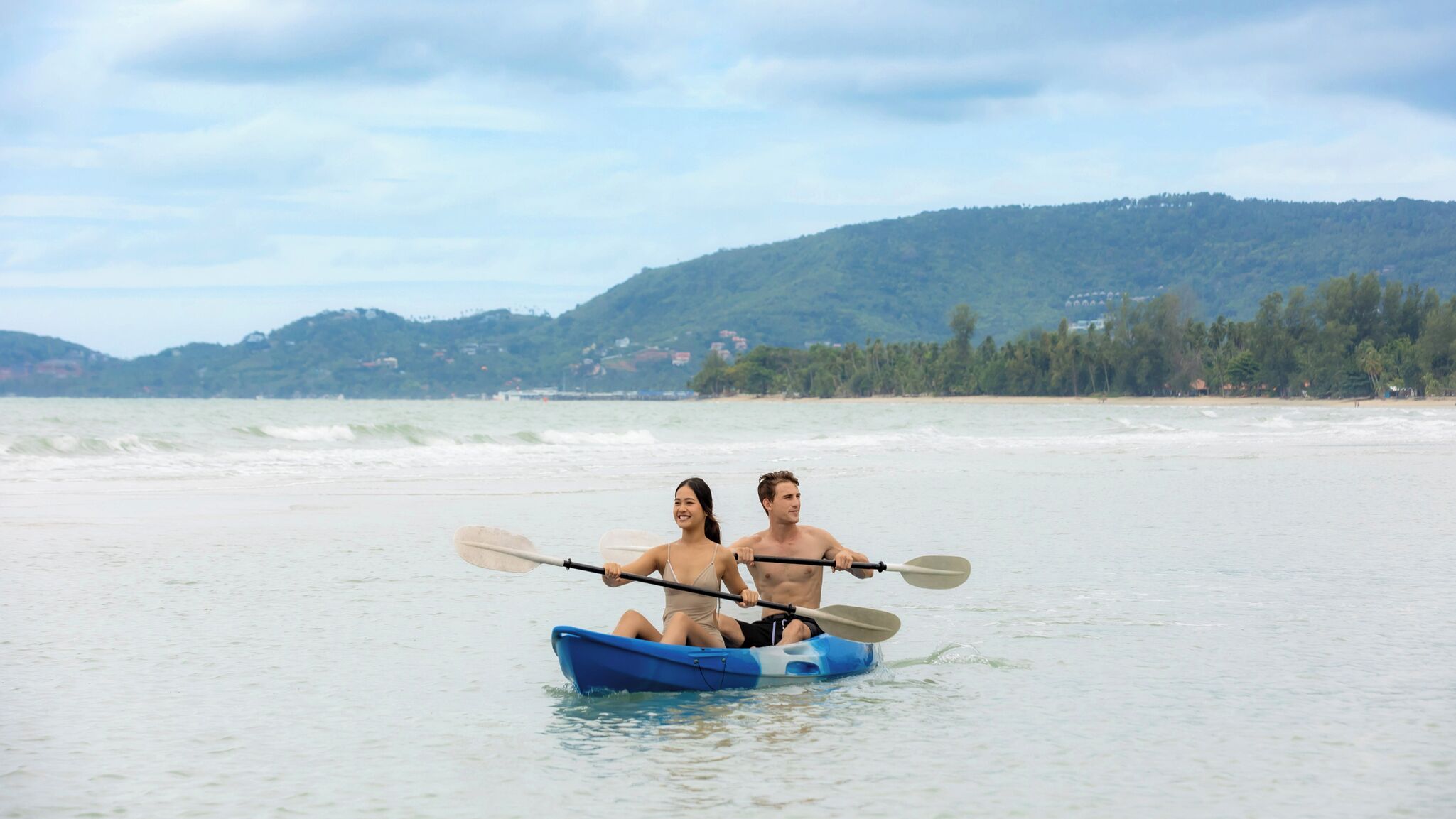 Una pareja en un kayak azul sobre un mar tranquilo, con un paisaje montañoso al fondo.