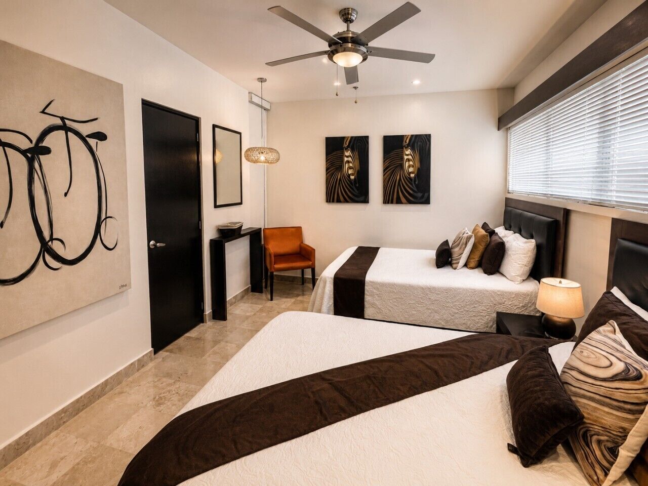 Une chambre d’hôtel avec deux lits, un ventilateur de plafond, des œuvres d’art accrochées aux murs, une chaise et une table de chevet avec une lampe.
