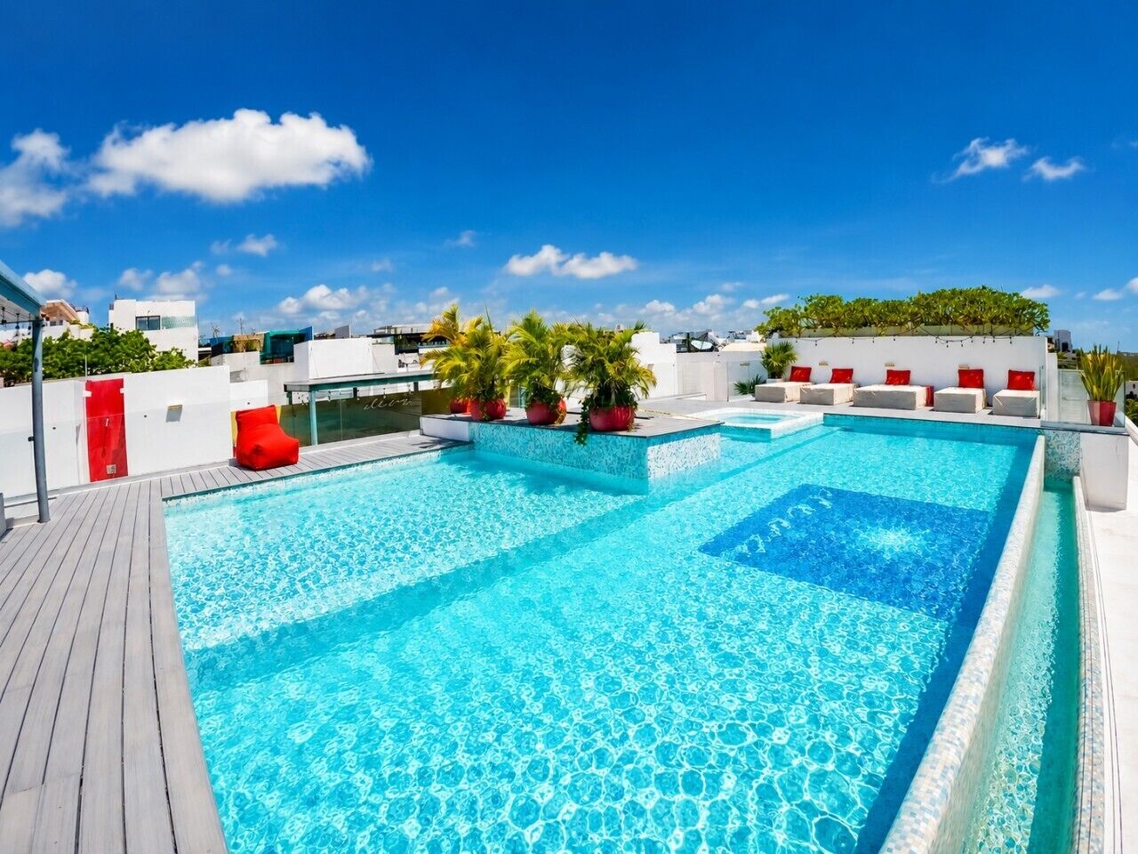 Une piscine sur le toit, dotée d’une terrasse en bois, entourée de plantes en pot et de sièges rouges.