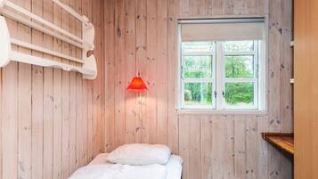 A room with wooden walls, a bed with white bedding, a red wall-mounted lamp, and a window with a view of greenery.