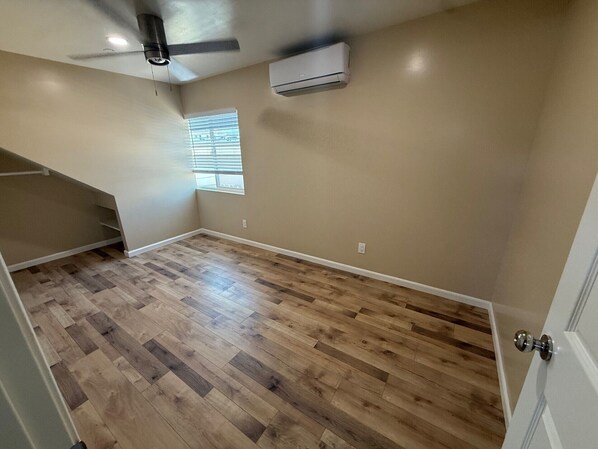 A room with wooden flooring, a ceiling fan, an air conditioning unit, and a window with blinds.