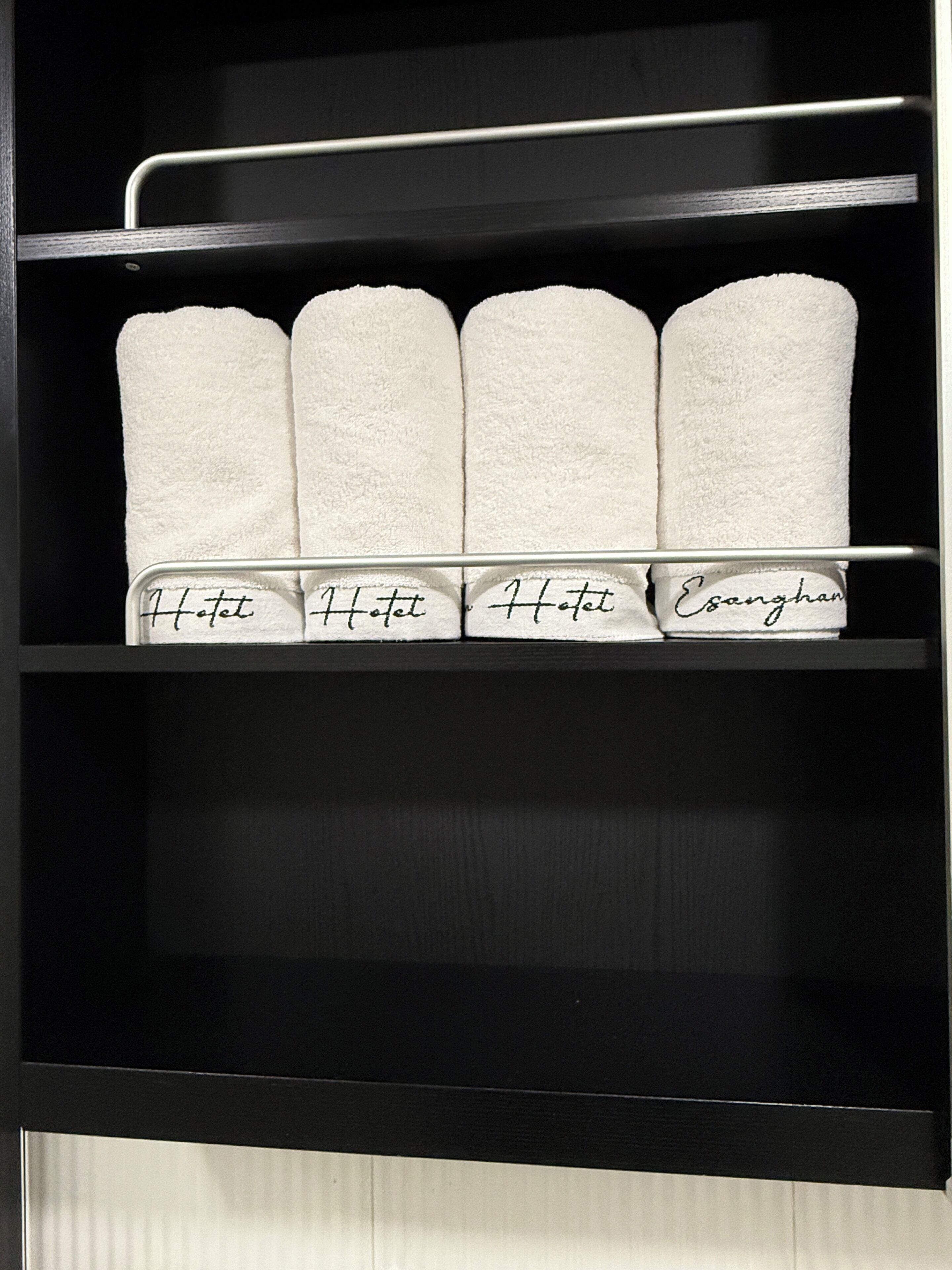 A hotel room with a black shelf holding four white towels and a metal rack above them.