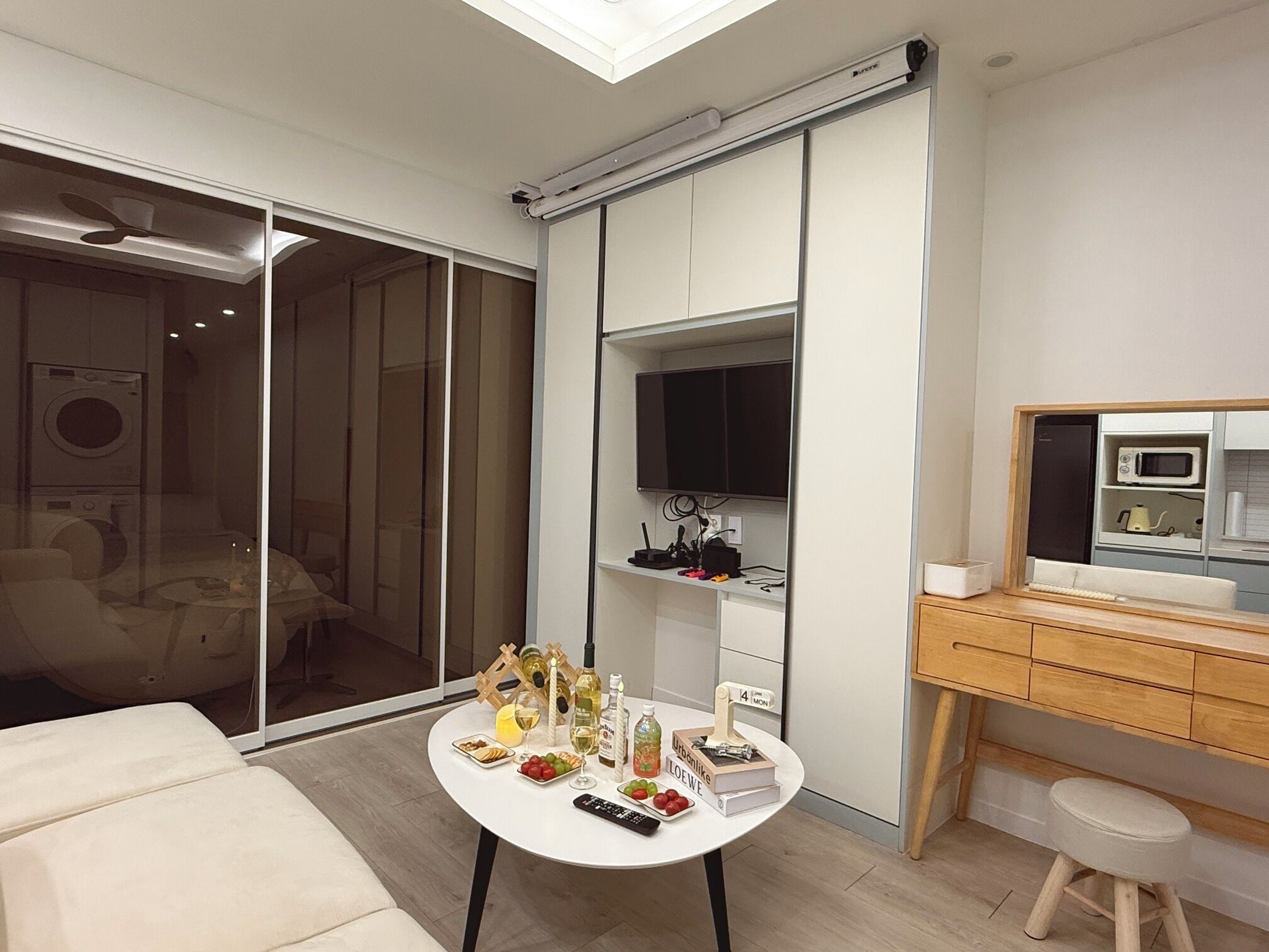 A modern living room with a sliding glass door leading to a laundry area, a round table with a remote control, and a small stool.