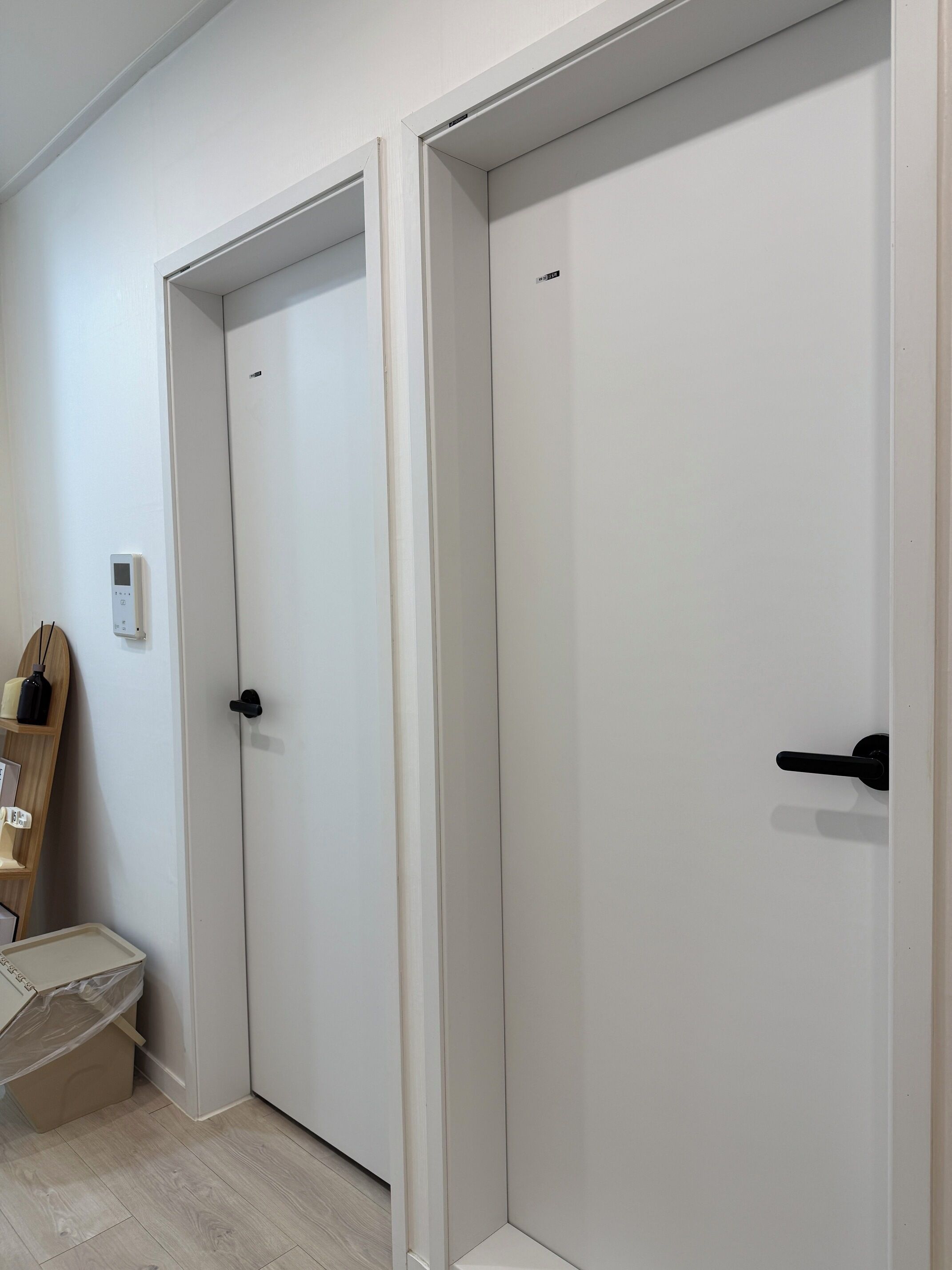 Un pasillo con dos puertas cerradas, un pequeño cesto de basura y un interruptor de luz en la pared.