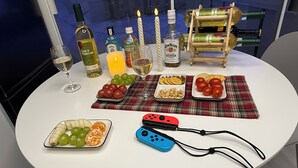 A table with various bottles, glasses, snacks, and a Nintendo Switch on a plaid placemat.