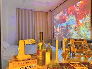 A hotel room with a bed, a table with a bottle, candles, a wine glass, and a projector screen displaying a Christmas tree.