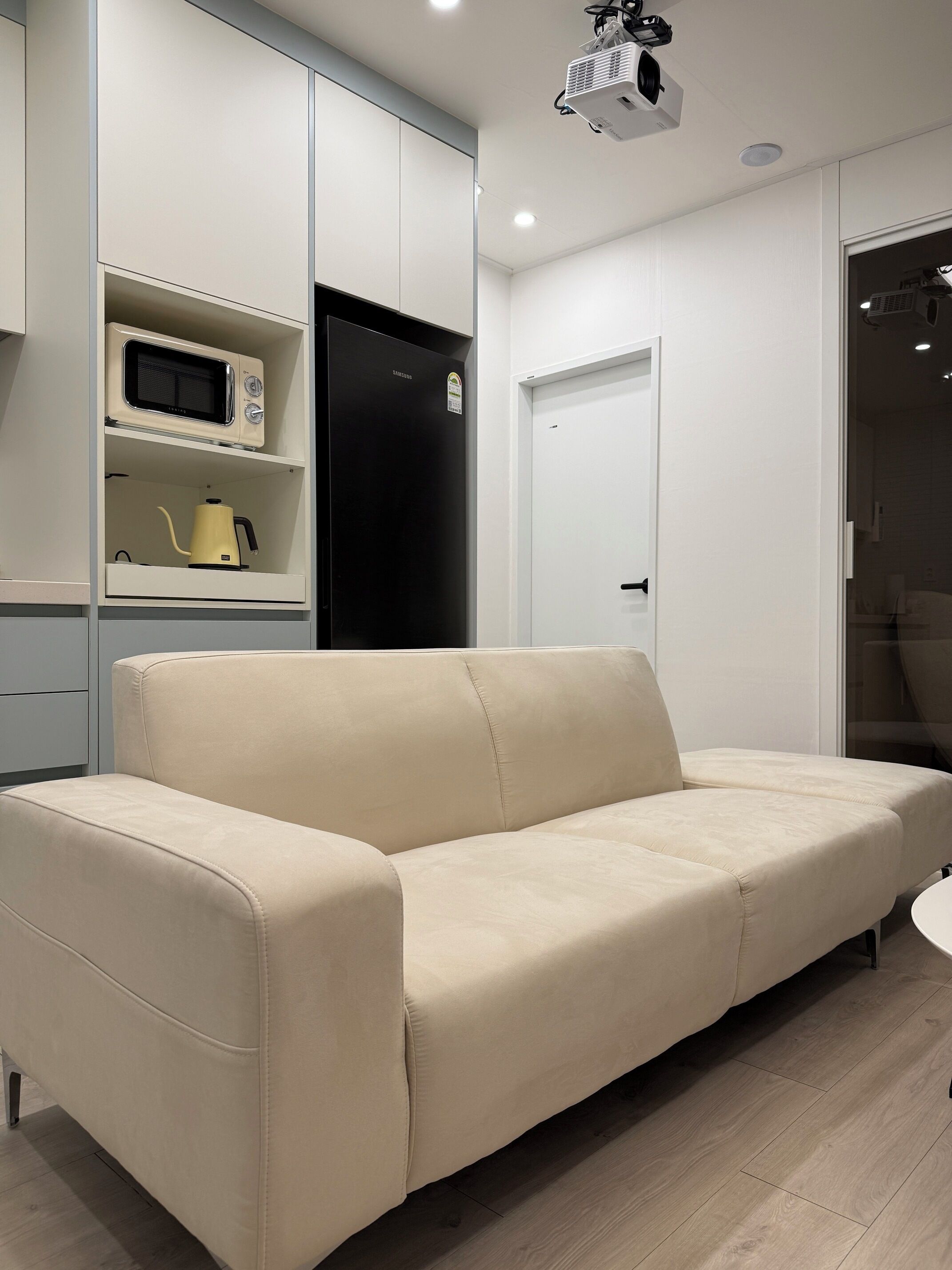 Una sala moderna con un sofá color beige, un microondas, un hervidor y un refrigerador negro.