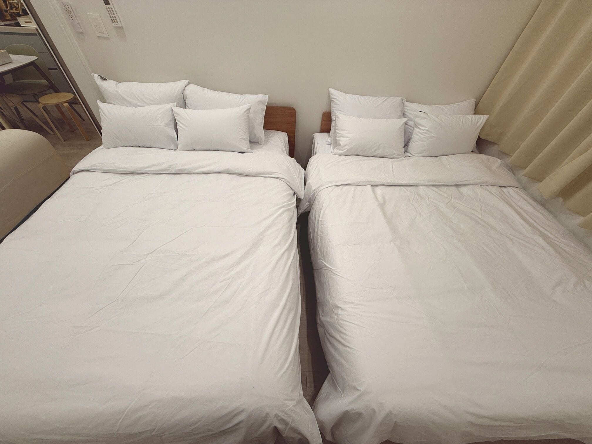 Deux lits simples avec du linge de lit et des oreillers blancs, des têtes de lit en bois, et une petite table avec des chaises à l’arrière-plan.