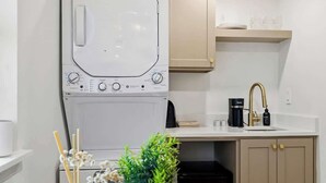 Una cocina compacta con lavadora y secadora integradas, un lavamanos con grifo dorado y una mesa redonda de comedor con planta en maceta.