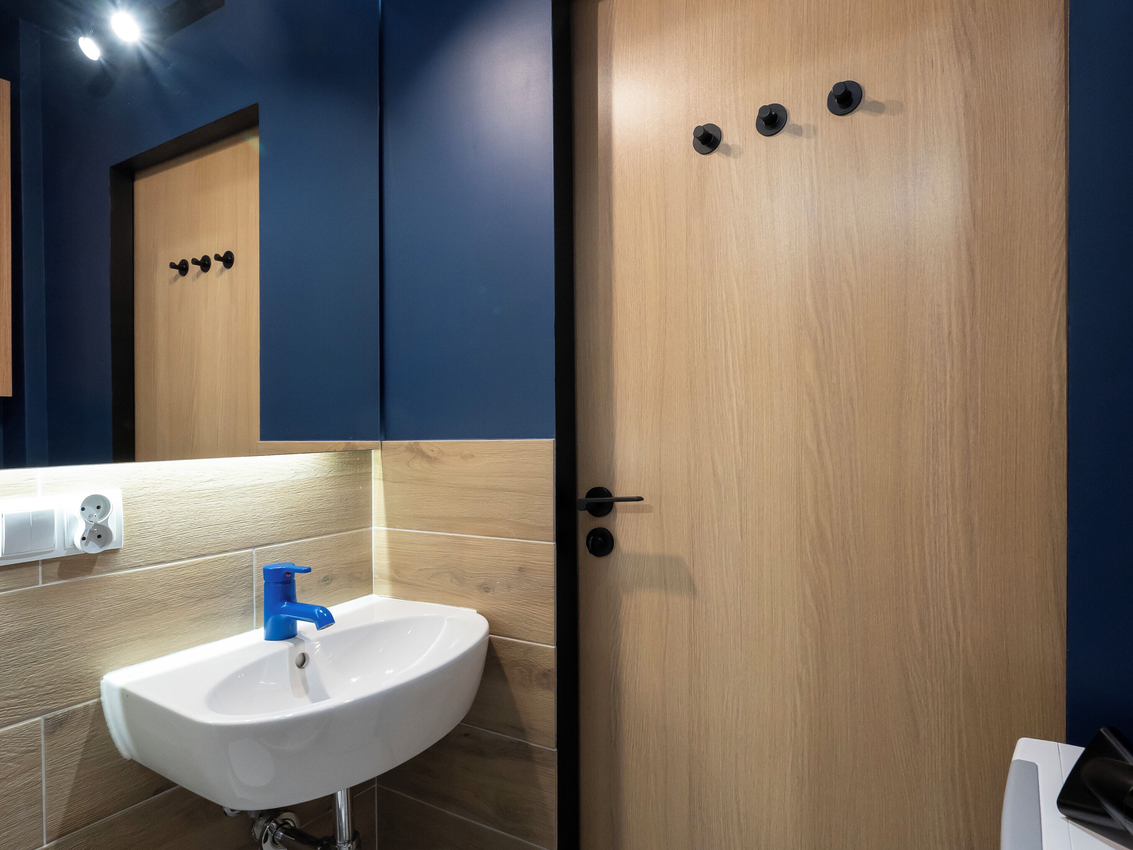 Un baño con un lavamanos blanco, un grifo azul, una puerta de madera con tres pomos negros y un espejo sobre el lavamanos.