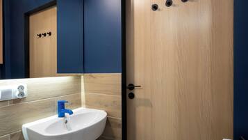 Un baño con un lavamanos blanco, un grifo azul, una puerta de madera con tres pomos negros y un espejo sobre el lavamanos.