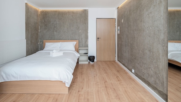 Ett modernt hotellrum med en säng i trä, ett litet nattduksbord, en papperskorg och en dörr.