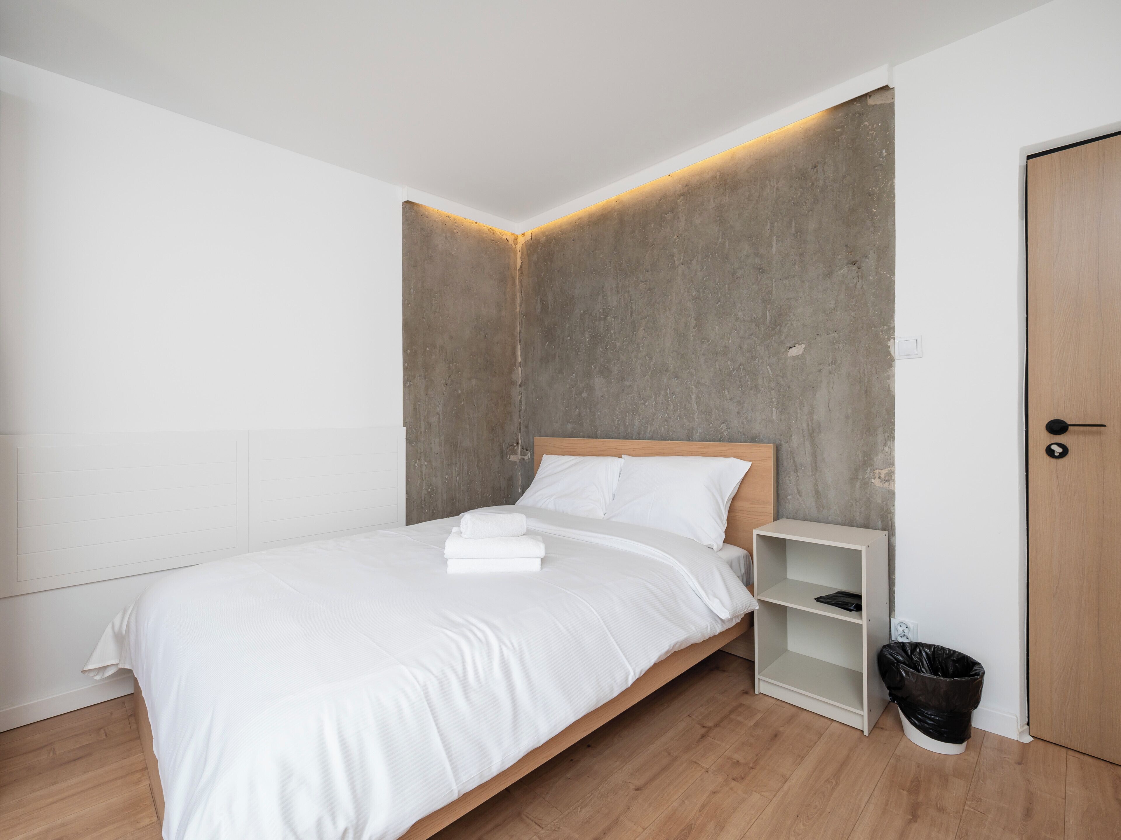 A modern bedroom with a wooden bed, white linens, a concrete accent wall, a white nightstand, and a wooden door.