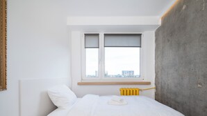 Ein Schlafzimmer mit Bett, Fenster mit Jalousien, Heizkörper und einer Betonwand.