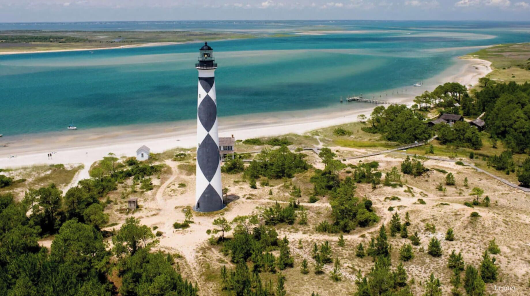 Un phare à l’architecture distinctive, ornée d’un motif en losanges, entouré d’un paysage côtier comprenant une plage, une jetée et quelques arbres dispersés.