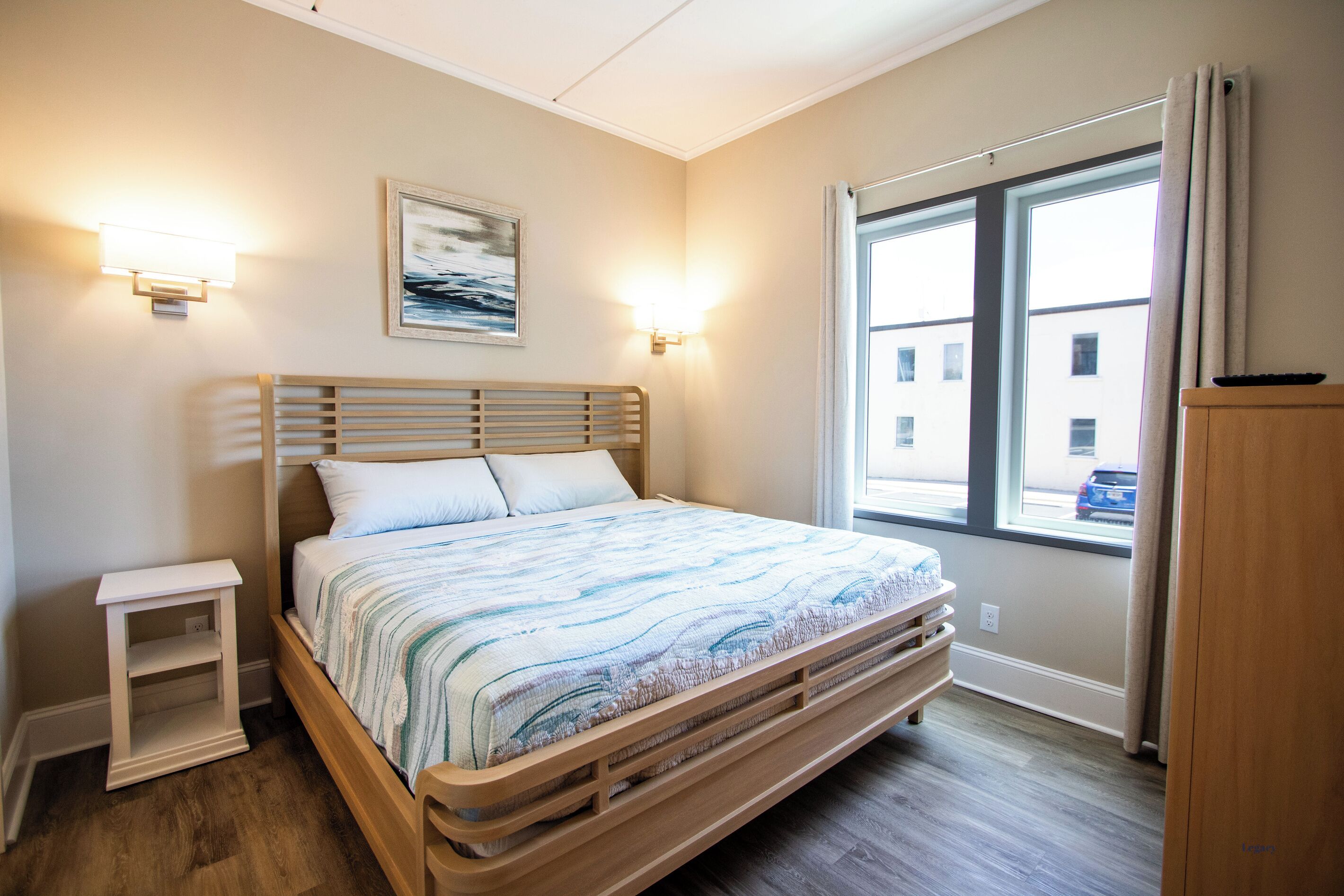 Une chambre à coucher avec un lit en bois, une table de chevet, une lampe fixée au mur et une fenêtre donnant sur des immeubles.