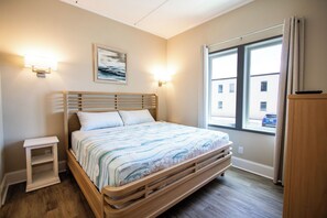 A bedroom with a wooden bed, a nightstand, a wall-mounted light, and a window with a view of buildings.