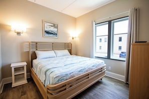 A bedroom with a wooden bed, bedside lamps, a nightstand, a window with a view, and a painting on the wall.