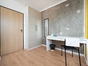 Una habitación con piso de madera, un escritorio blanco, una silla negra, un armario blanco y una pared de concreto.