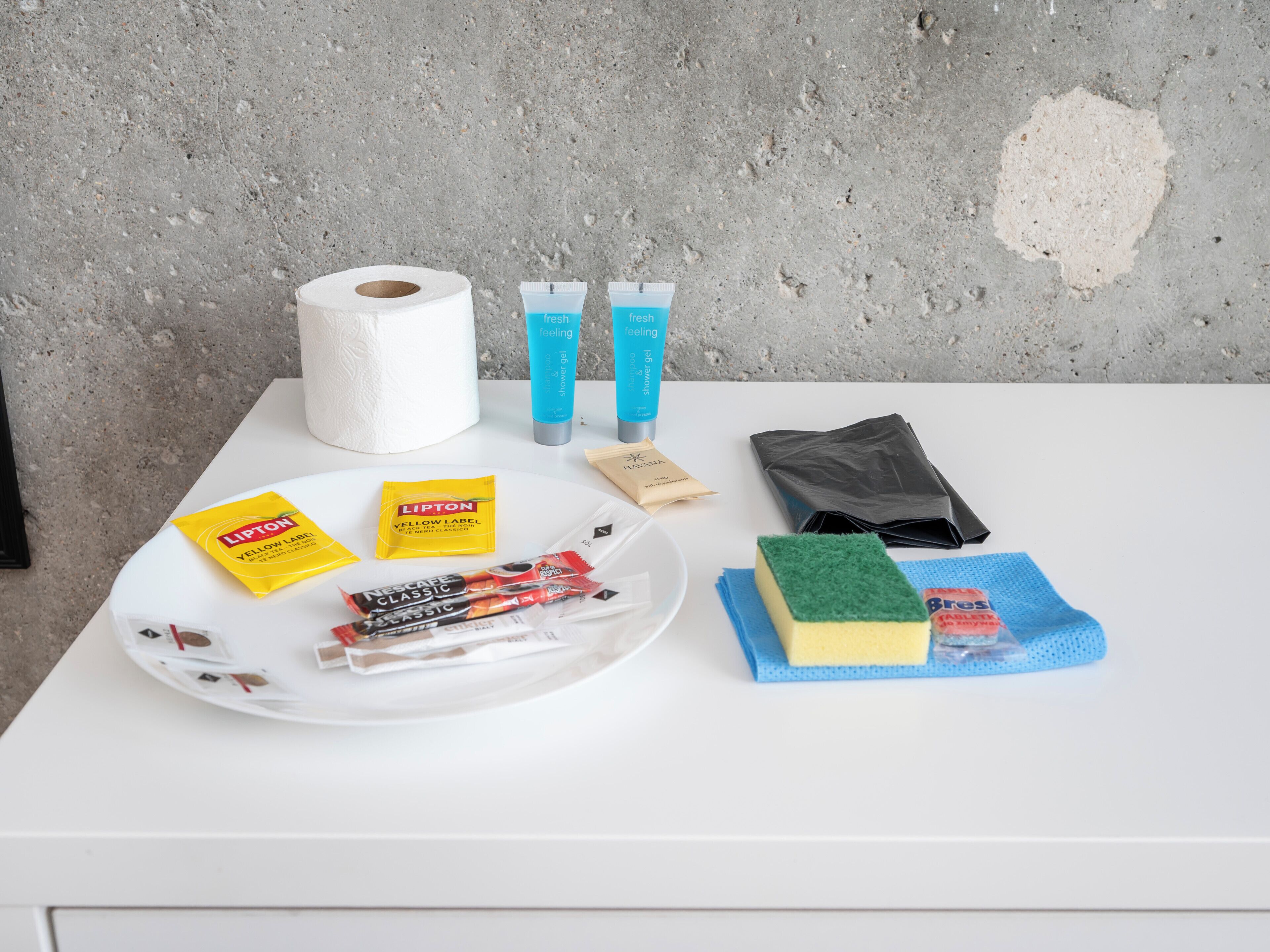 Una mesa con artículos de aseo, incluyendo un rollo de papel higiénico, gel de ducha y un paquete de café Nescafe Classic.