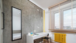 A modern room with a white desk, a black chair, a large mirror, and a window with blinds. - Lermontowa Comfort Room #3 (Łódź)