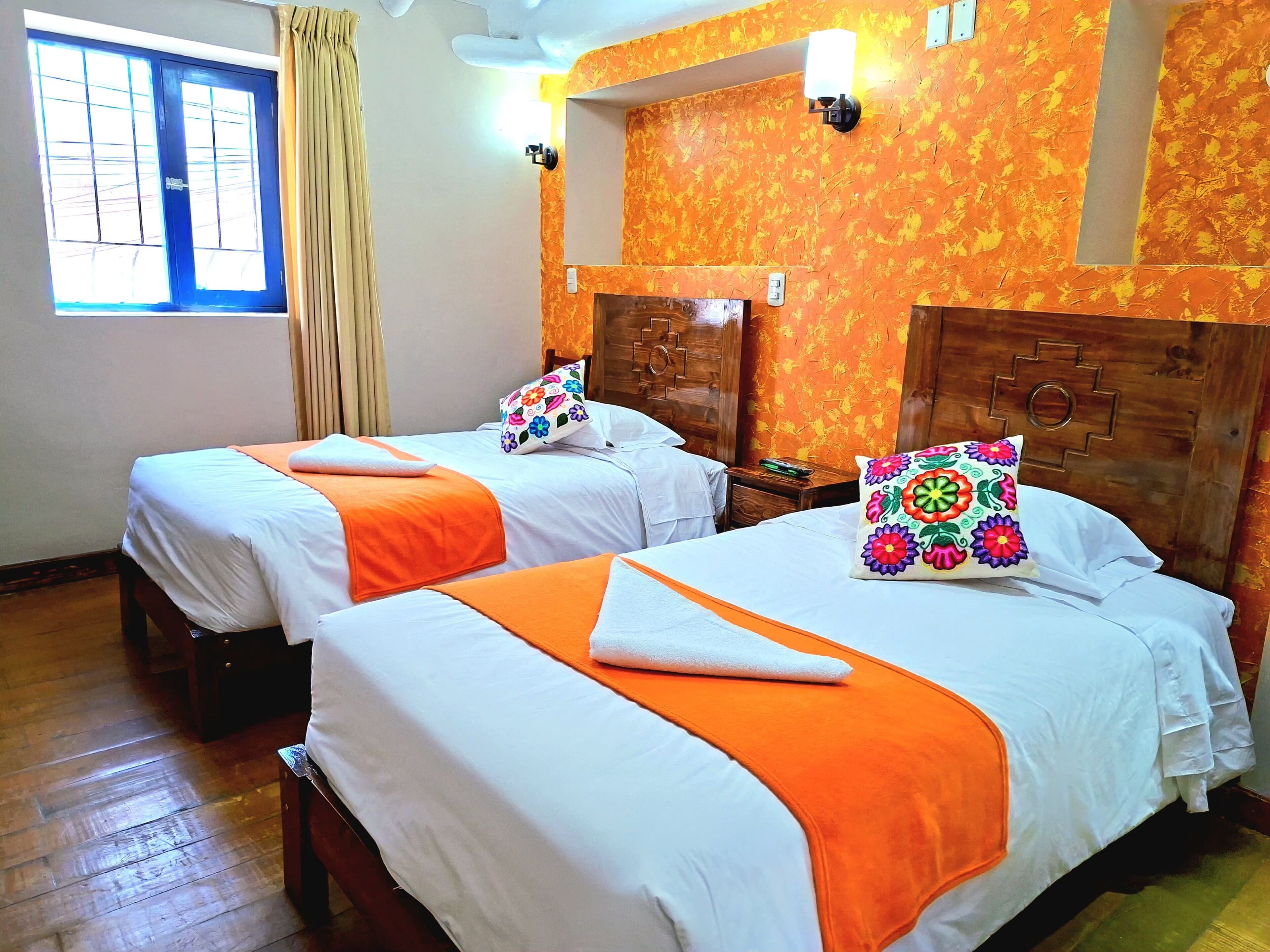 Dos camas con ropa de cama blanca y naranja, un cabecero de madera con un espejo, una ventana con cortinas y una pared con estampado floral.