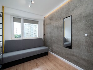 A modern room with a grey sofa, a large window, a concrete accent wall, and a mirror. - Lermontowa Comfort Room #5 (Łódź)