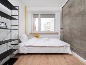 A small, modern bedroom with a bed, a black metal shelf unit, a window with a view of buildings, and a radiator. - Lermontowa Comfort Room #5 (Łódź)
