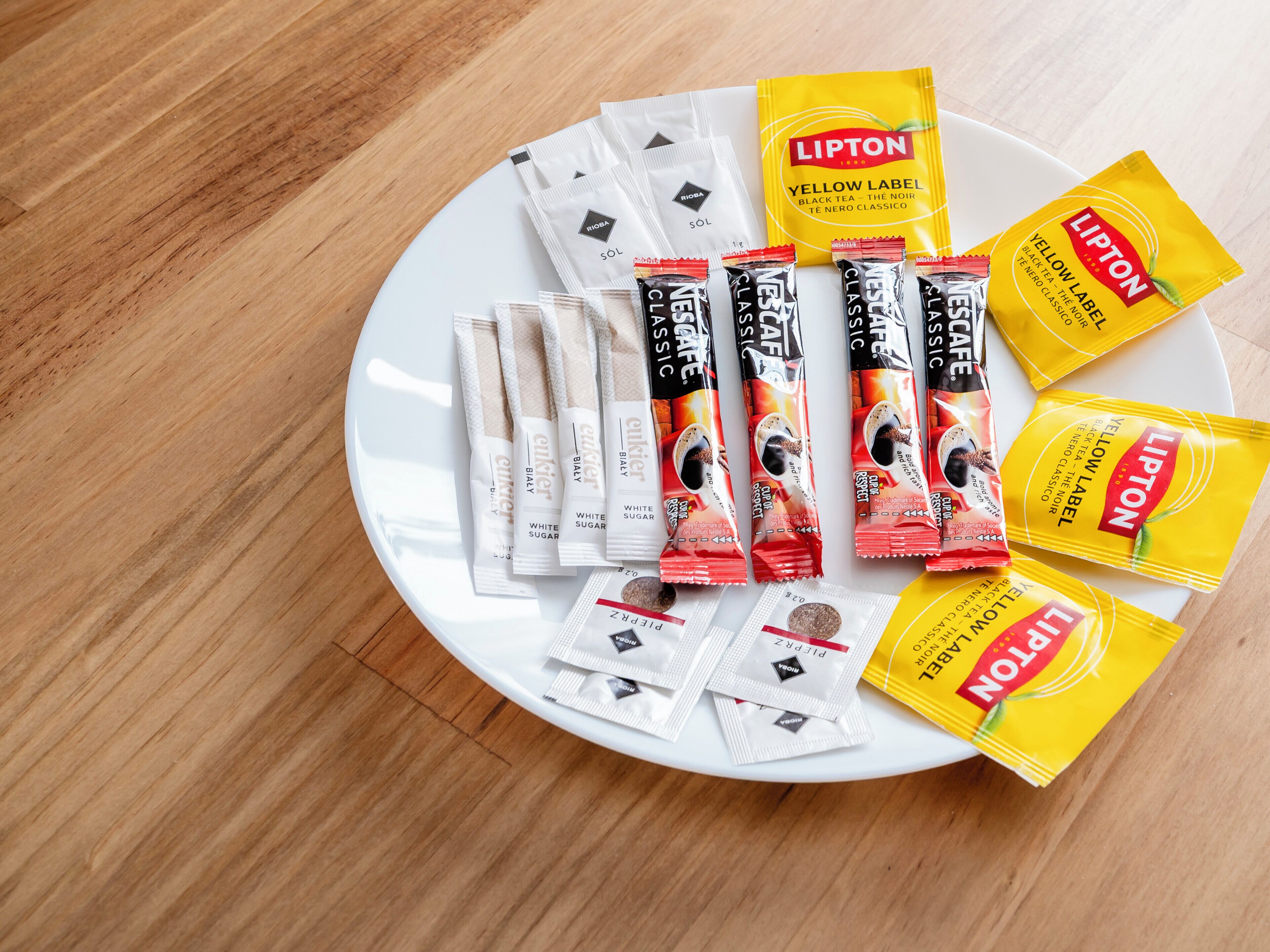 A white plate with various tea packets and sachets, including Lipton and Rioha brands, arranged on a wooden surface.
