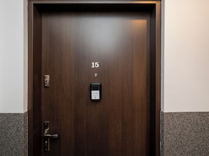 Uma porta de quarto de hotel em madeira escura, com um cadeado de teclado numérico, um interfone e uma pequena placa acima da porta.