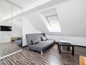 Ein modernes Schlafzimmer im Loft-Stil mit schrägverdeckter Decke, einem grauen Sofa, einem kleinen Tisch und einem an der Wand befestigten Fernseher.
