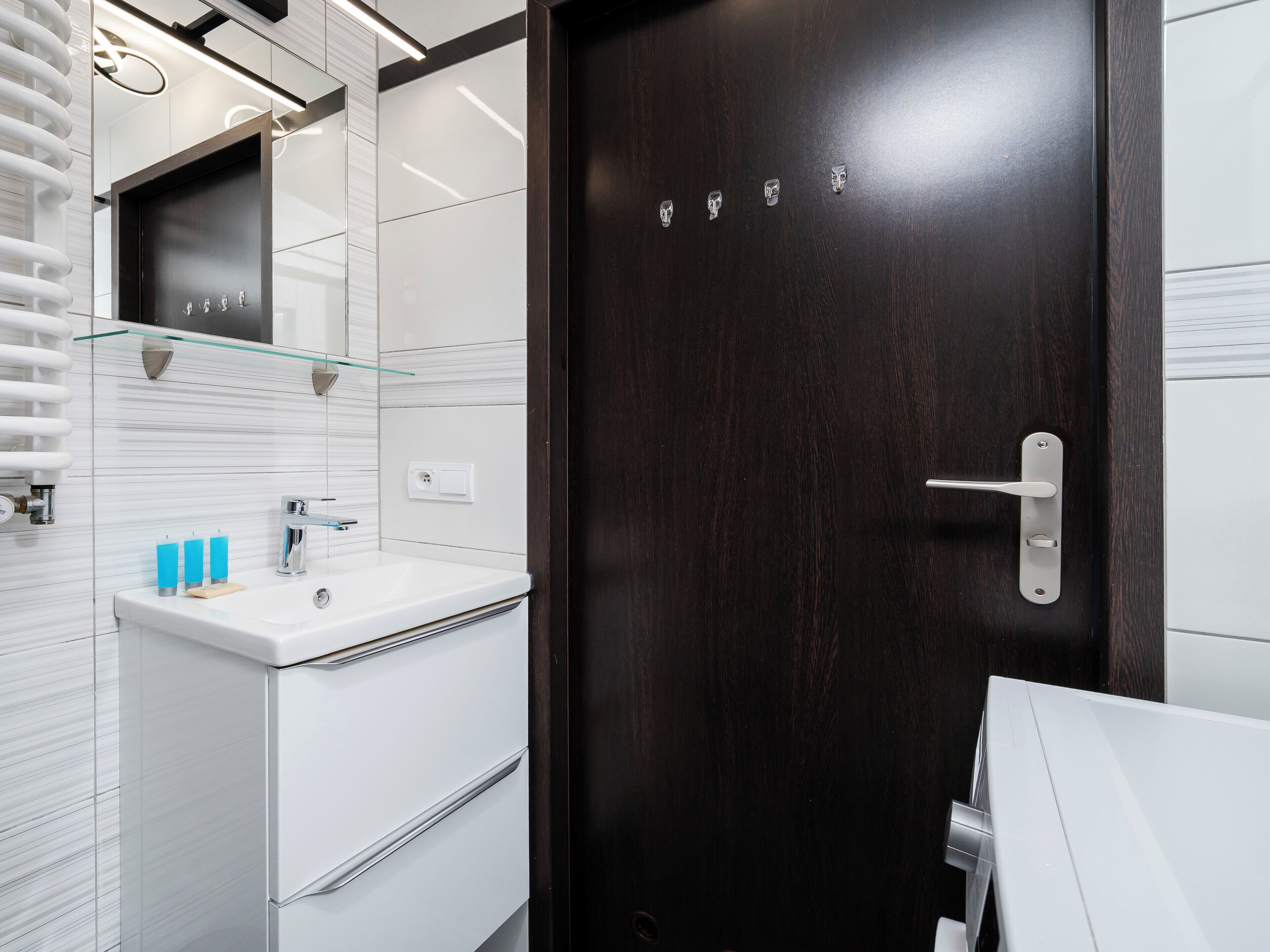 Ein modernes Badezimmer mit einer dunkelholzigen Tür, weißen Fliesen, einer Glasablage und einem weißen Waschbecken.