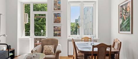 A room with a dining table, chairs, a sofa, and a coffee table. There are large windows with curtains, a radiator, and a framed picture on the wall.