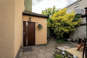 Ein Gebäude mit einer braunen Tür, einem kleinen Garten mit einem Baum und einer gelb gemusterten Matte.