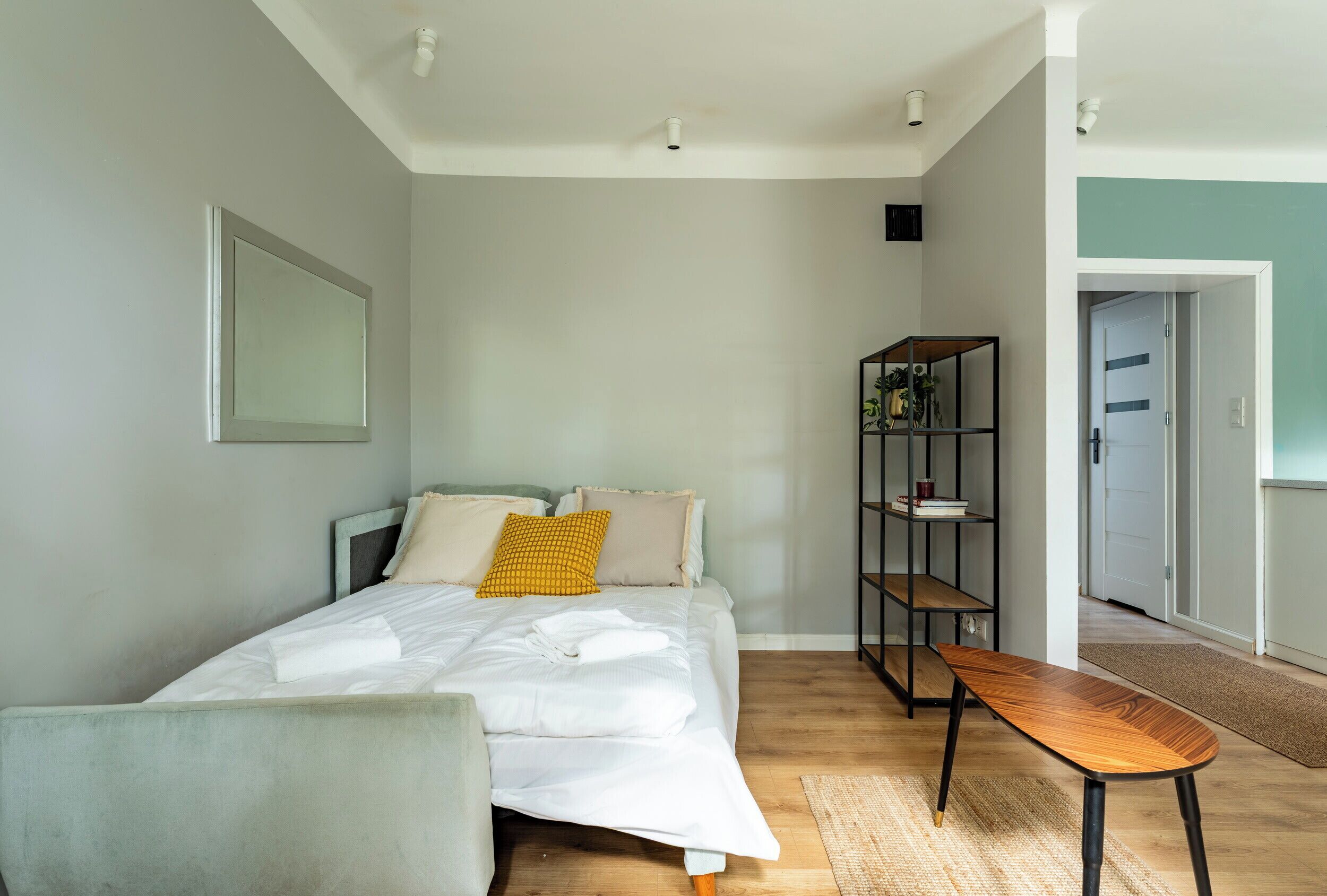 Een slaapkamer met een bed, nachtkastje, stoel, plank met boeken, en een deur naar een andere kamer.