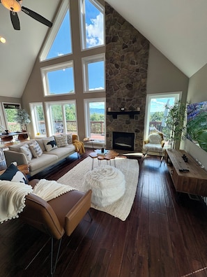 A spacious living room with a stone fireplace, large windows, and a high ceiling.
