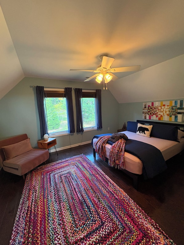 A bedroom with a bed, a chair, a ceiling fan, a window with curtains, and a colorful rug.