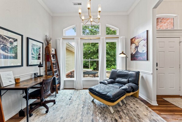 A room with a desk, chair, black leather sofa, large windows, and artwork.