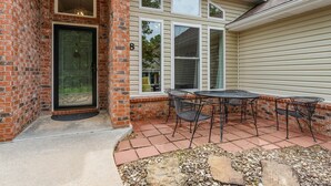 Una entrada de ladrillo con puerta de cristal, un patio con mesa y sillas, y una casa con varias ventanas.