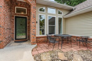A brick entrance with a glass door, a patio with a table and chairs, and a house with multiple windows.