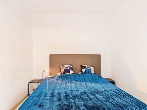 Ein Bett mit blauer, gemusterter Bettdecke, zwei Kissen mit abstrakten Mustern, ein Nachttisch mit Lampe und eine schlichte weiße Wand.