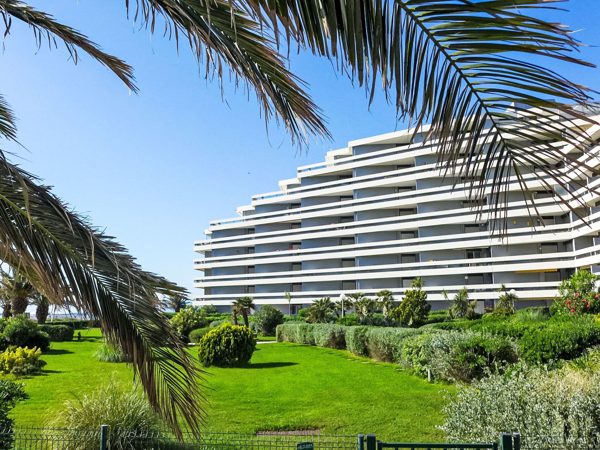 A modern multi-story hotel with a flat roof, surrounded by well-maintained greenery and a clear blue sky.