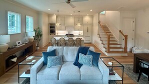 A spacious living area with a sofa, coffee table, and a dining table. There is a kitchen in the background with a staircase leading to another level. - King & Queen- All 5 Units (Auburn)